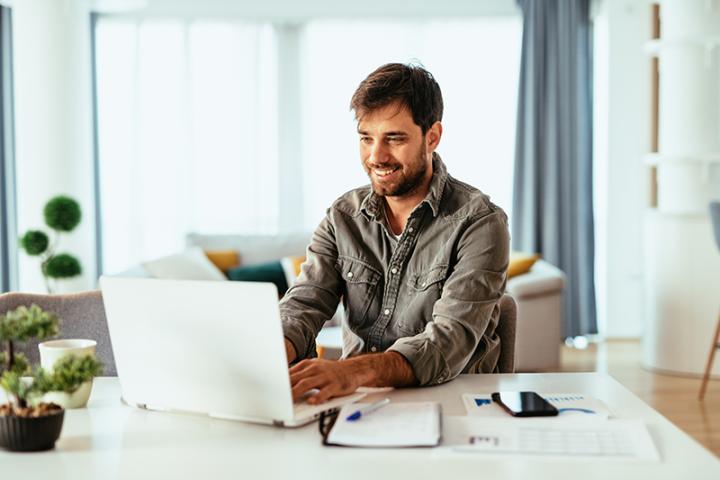 A man sits at a laptop smiling in a well-lit room.